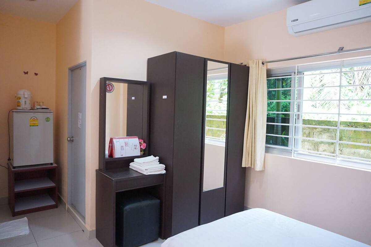 Un dormitorio con cama, armario, espejo, un escritorio pequeño con espejo, calentador y ventana con vistas a zonas verdes.