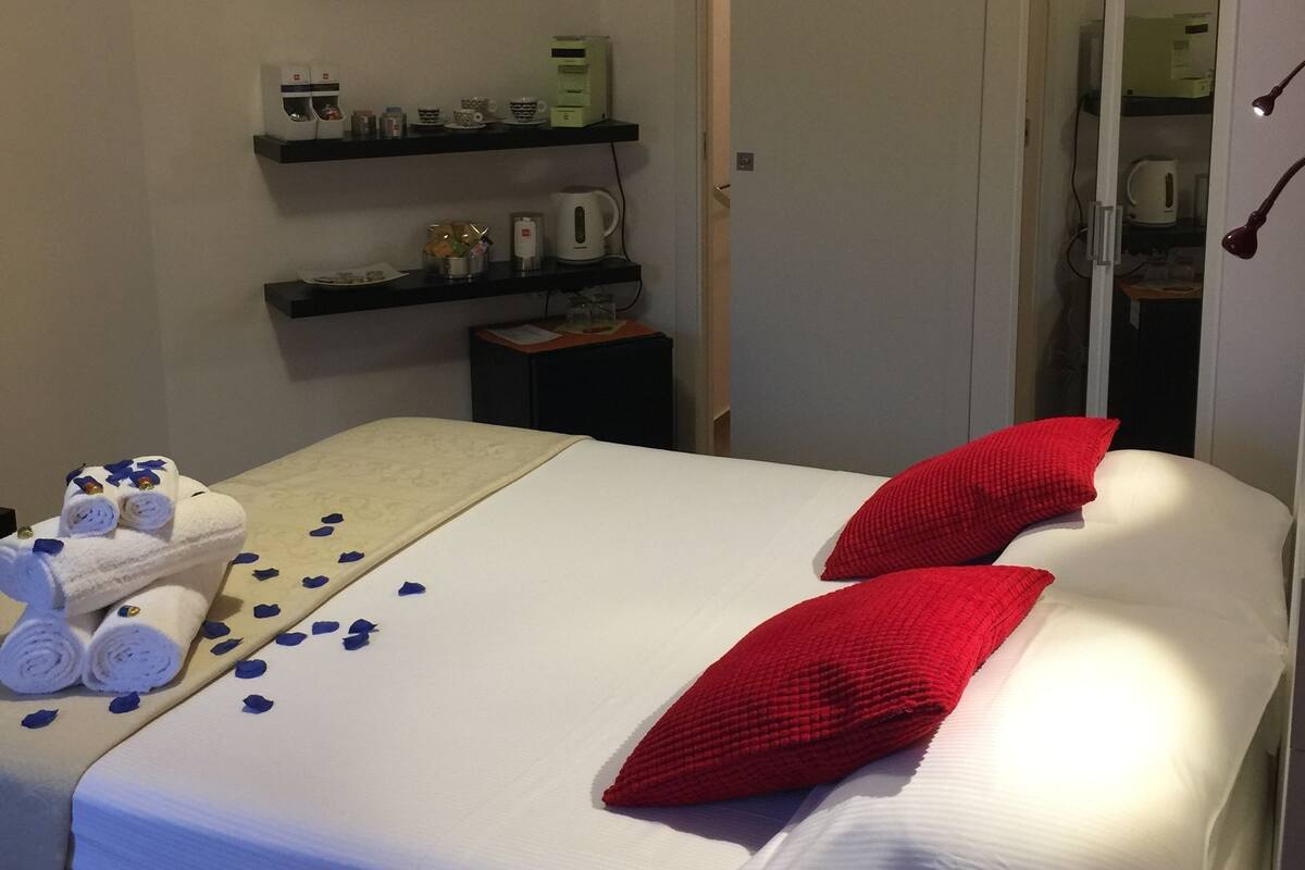 A hotel room with a neatly made bed, a small bedside table with a lamp, a shelf with toiletries, and a framed artwork on the wall.