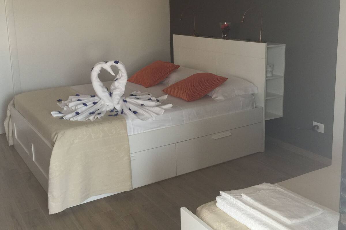A neatly made bed with a heart-shaped towel arrangement, a bedside table with a vase, and a wall-mounted shelf.