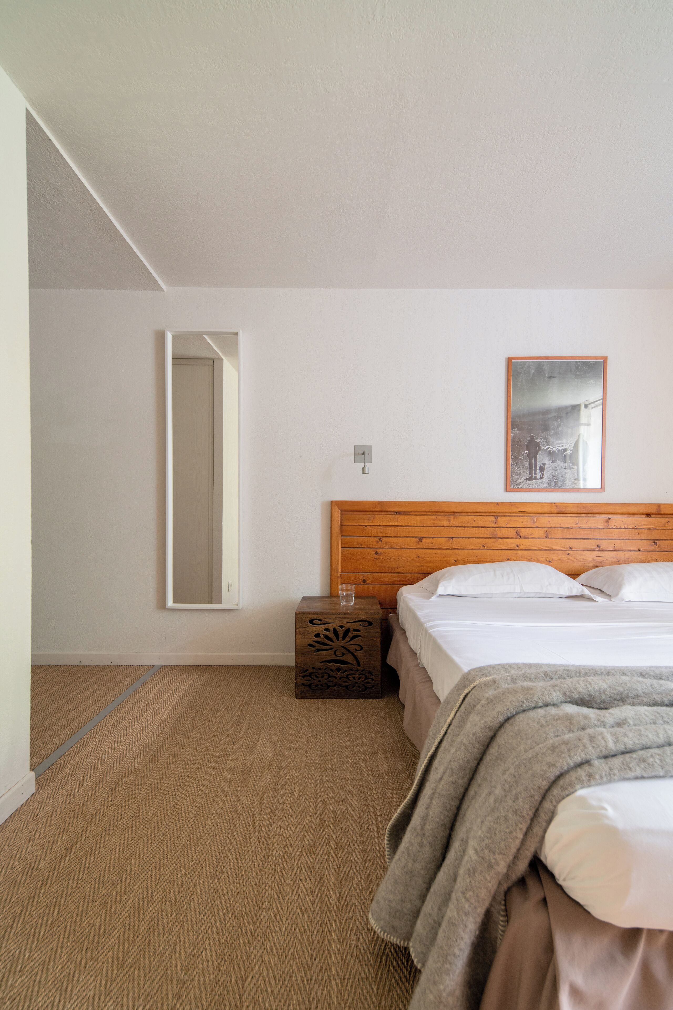 Een hotelkamer met een houten hoofdeinde, een bed met witte lakens, een spiegel, een lijst met een afbeelding aan de muur en een klein houten nachtkastje.