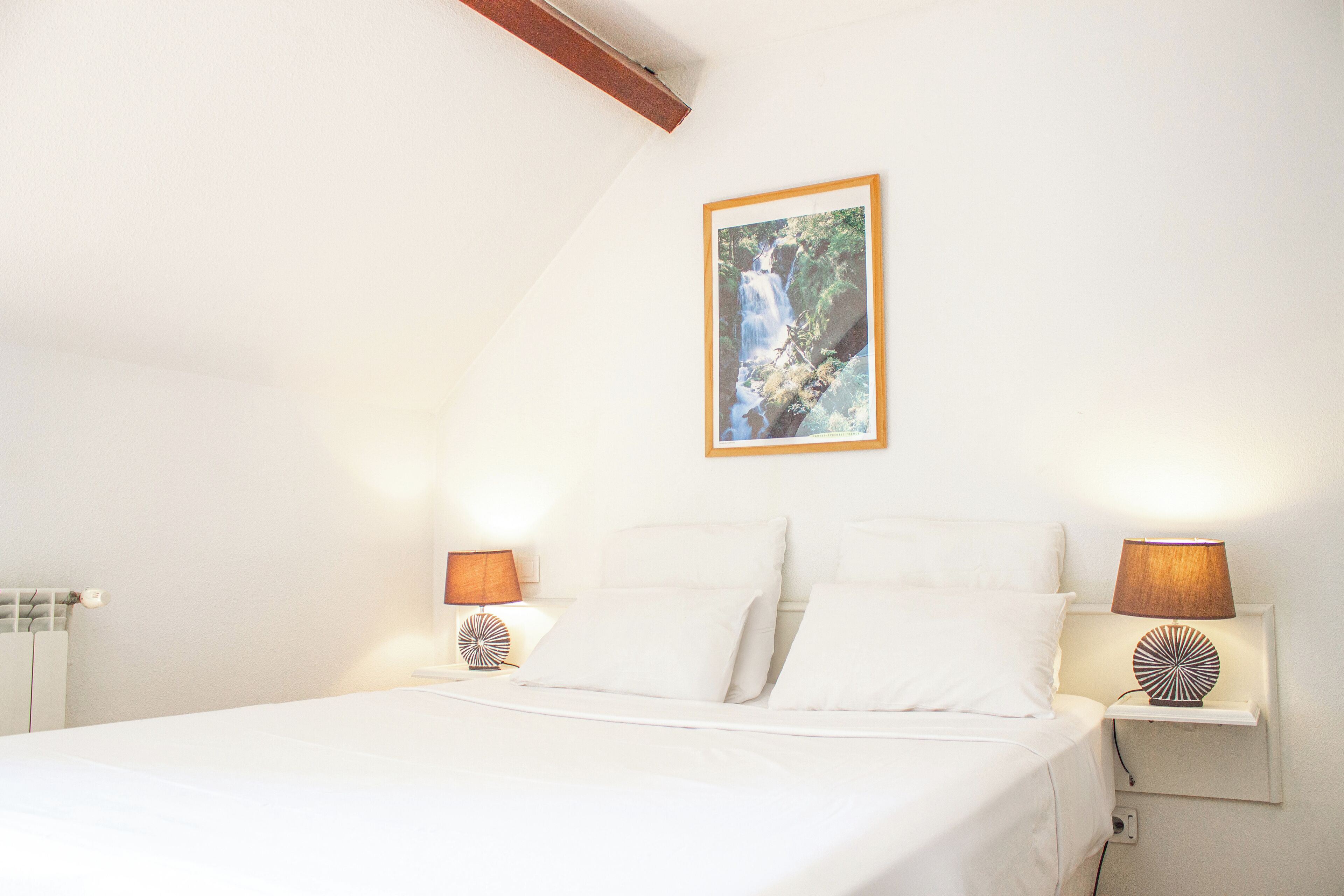 Une chambre à coucher avec un lit, deux lampes de chevet, un tableau accroché au mur et une petite étagère avec une lampe.