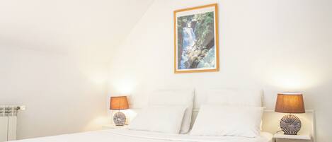 Un dormitorio con una cama, dos lámparas de noche, un cuadro colgado en la pared y una pequeña estantería con una lámpara.