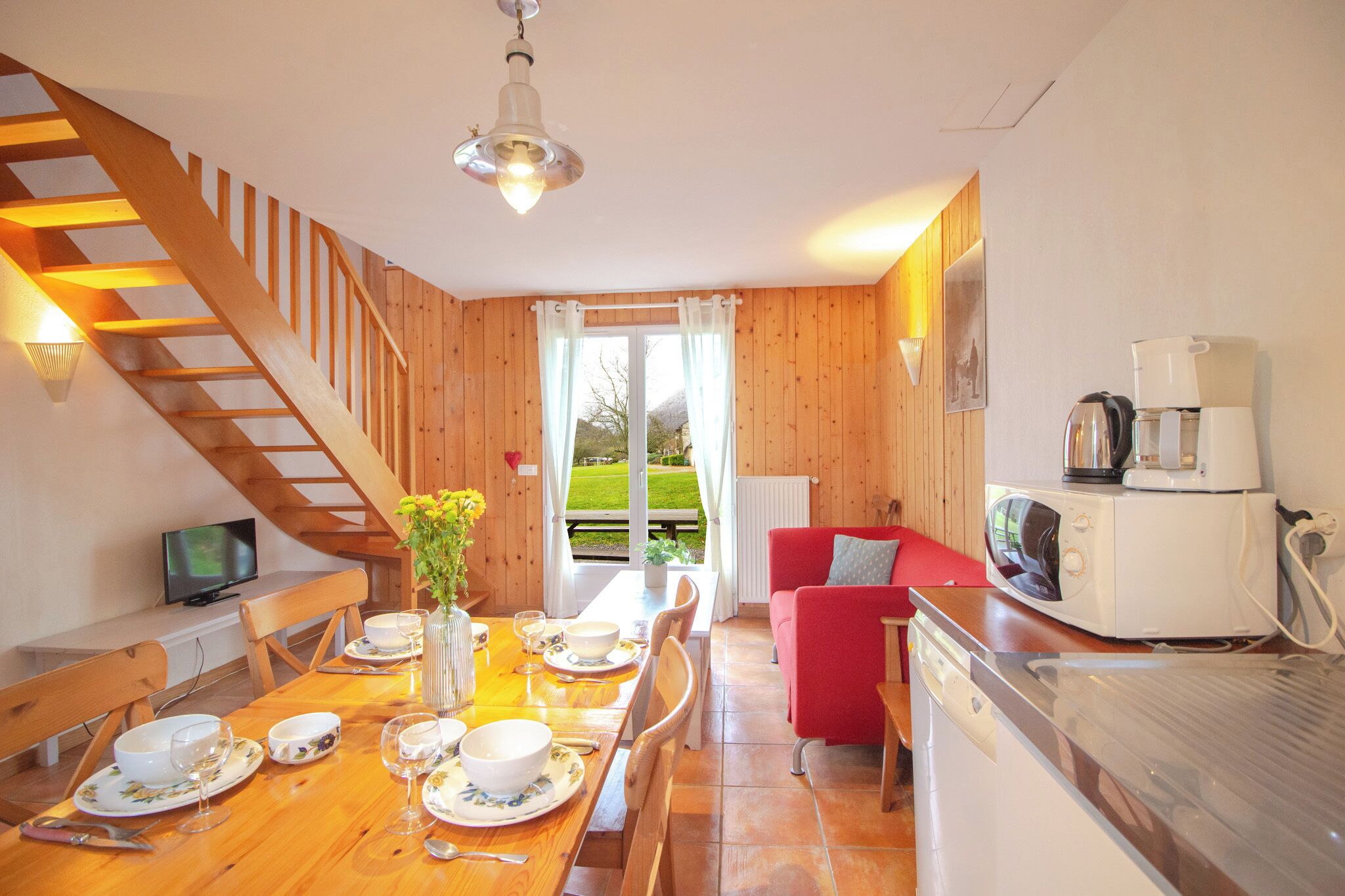 A dining area with a wooden table set for four, a red sofa, a kitchen area with a microwave, and a staircase leading to an upper level.