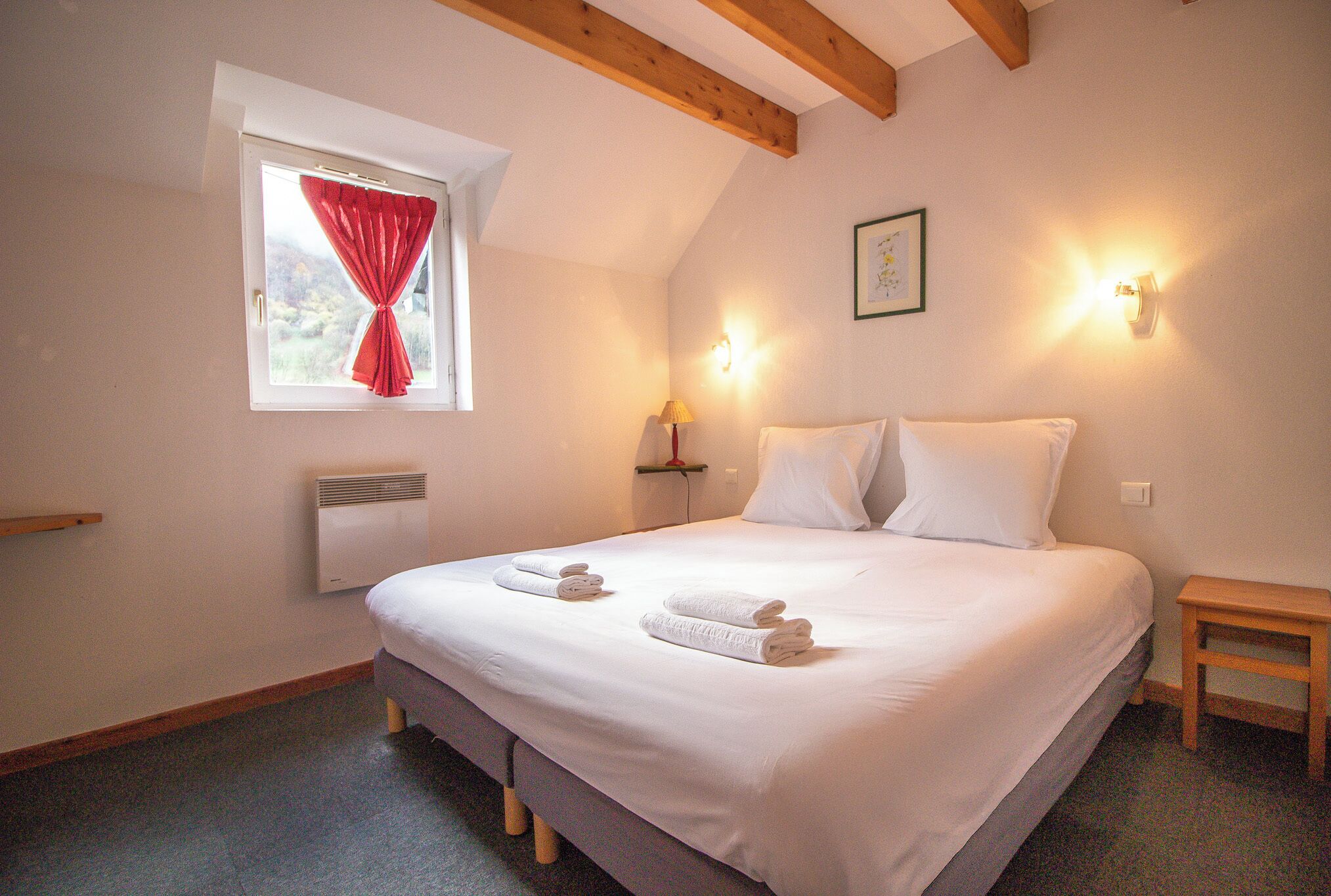 Un lit bien fait, avec du linge de lit blanc et deux serviettes, une fenêtre avec des rideaux rouges, une lampe de chevet et une petite table de nuit en bois.
