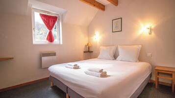 Un lit bien fait, avec du linge de lit blanc et deux serviettes, une fenêtre avec des rideaux rouges, une lampe de chevet et une petite table de nuit en bois.