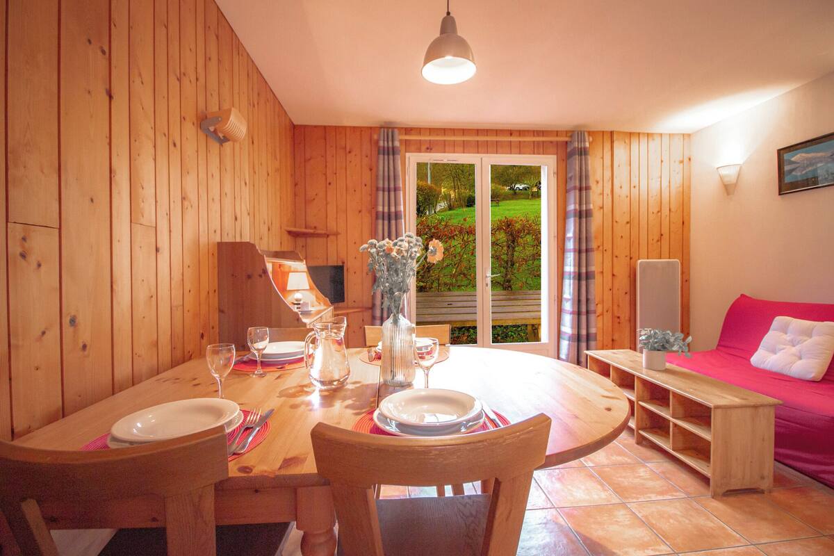 Un intérieur en bois avec une table à manger dressée pour deux personnes, un canapé rose et une vue sur un jardin, visible à travers une porte vitrée.