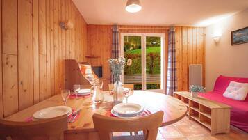 A wooden interior with a dining table set for two, a pink sofa, and a view of a garden through a glass door.