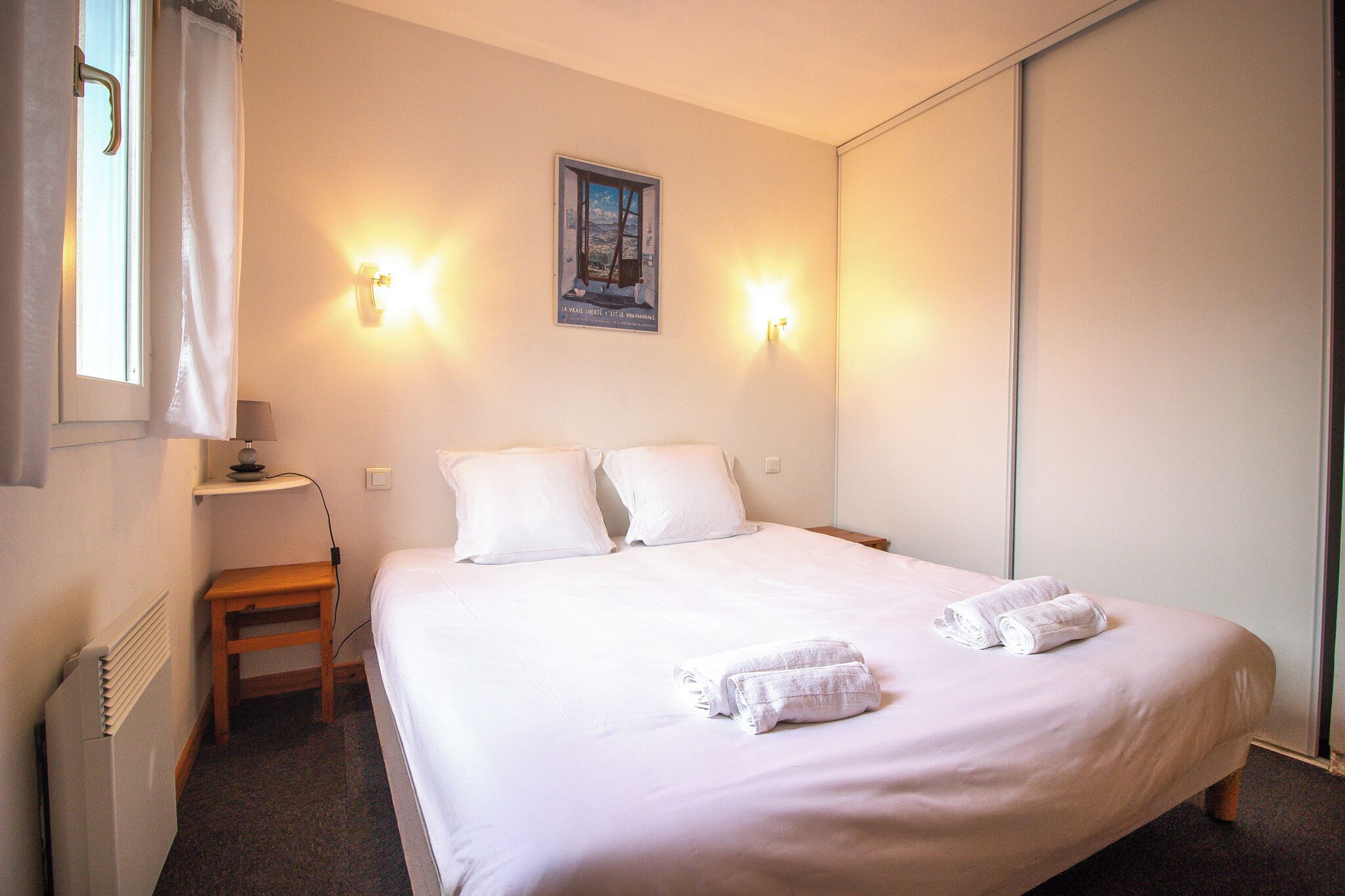A neatly made bed with white linens and towels, a bedside table with a lamp, and a wall-mounted picture.