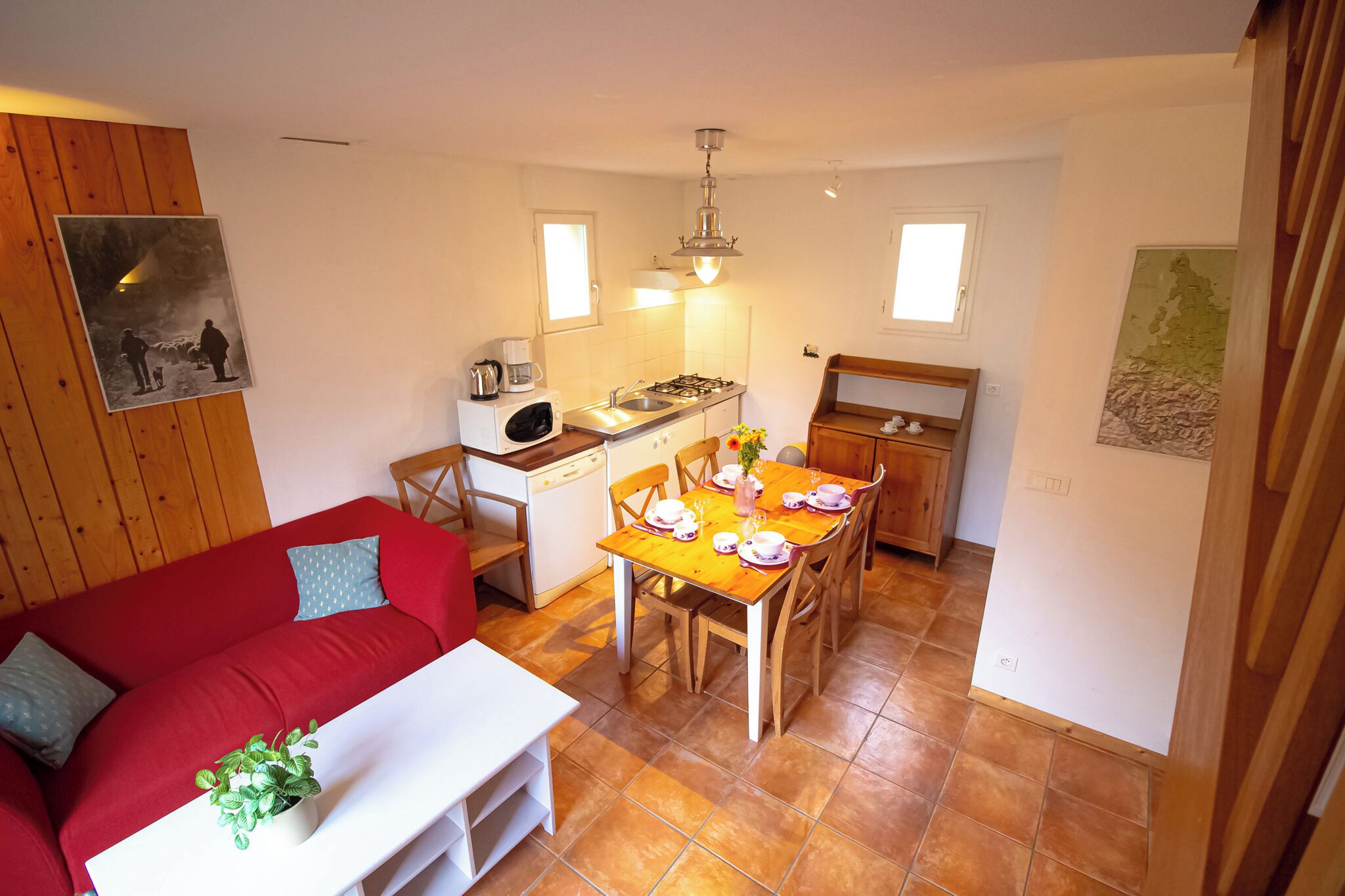 Un espacio habitable compacto con un sofá rojo, una mesa de comedor para cuatro, una zona de cocina con estufa y fregadero, y un pequeño armario blanco.