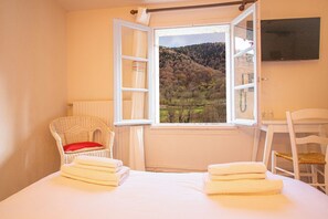 A room with a bed, a wicker chair with a red cushion, a white chair, and a window with a view of a forested hillside. - Garden & City Bagnères-de-Bigorre - Domaine de Ramonjuan (Bagnères-de-Bigorre)