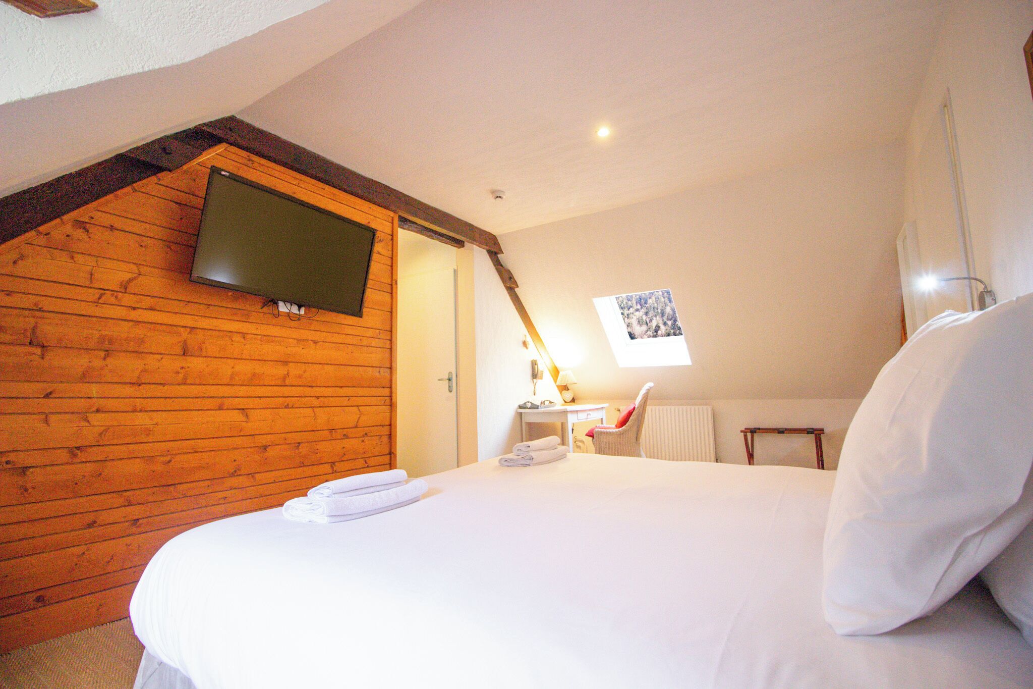 A bedroom with a wooden ceiling, a bed with white linens, a desk with a chair, and a TV mounted on the wall.