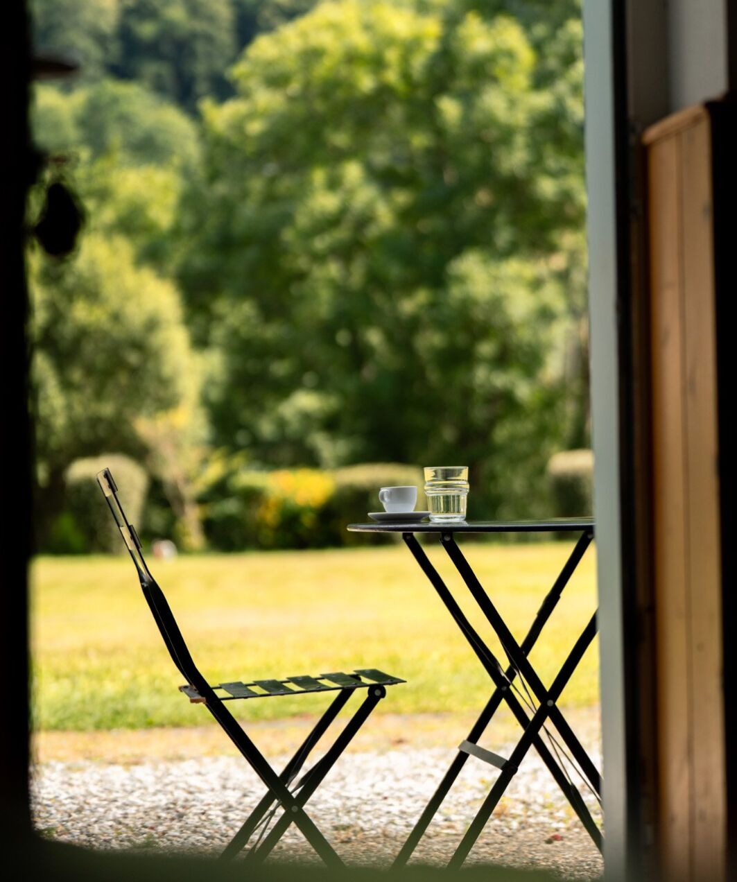 A table with a cup and glass, a chair, and a view of a green landscape through an open door.