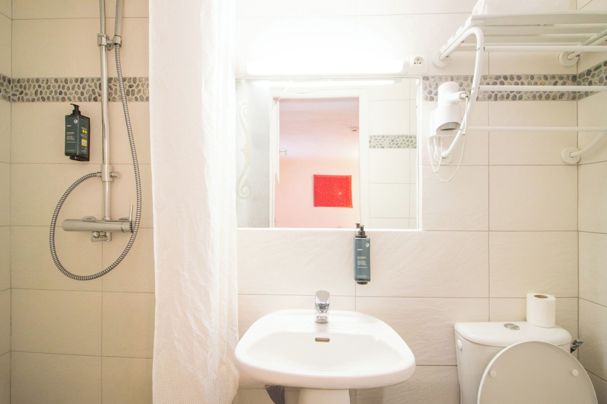 A bathroom with a white sink, toilet, and shower. There is a red object on the wall and a window above the sink.