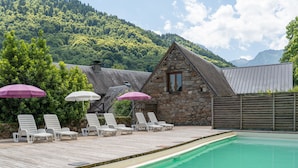 Zona de piscina con sillones de descanso y sombrillas, un edificio de piedra y montañas al fondo.