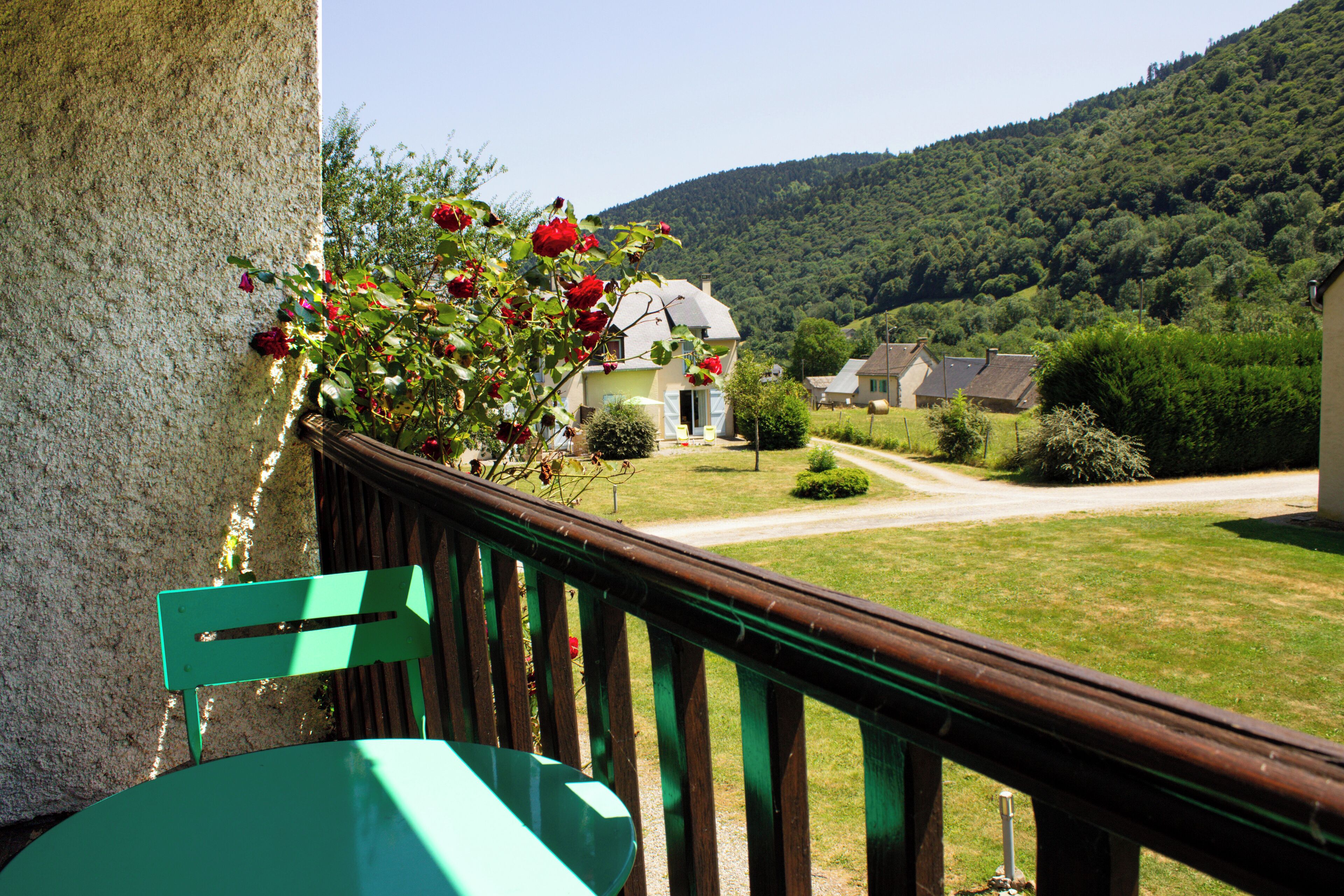 A balcony with a green chair, a flower box with red roses, and a view of a rural landscape with houses and a hill.