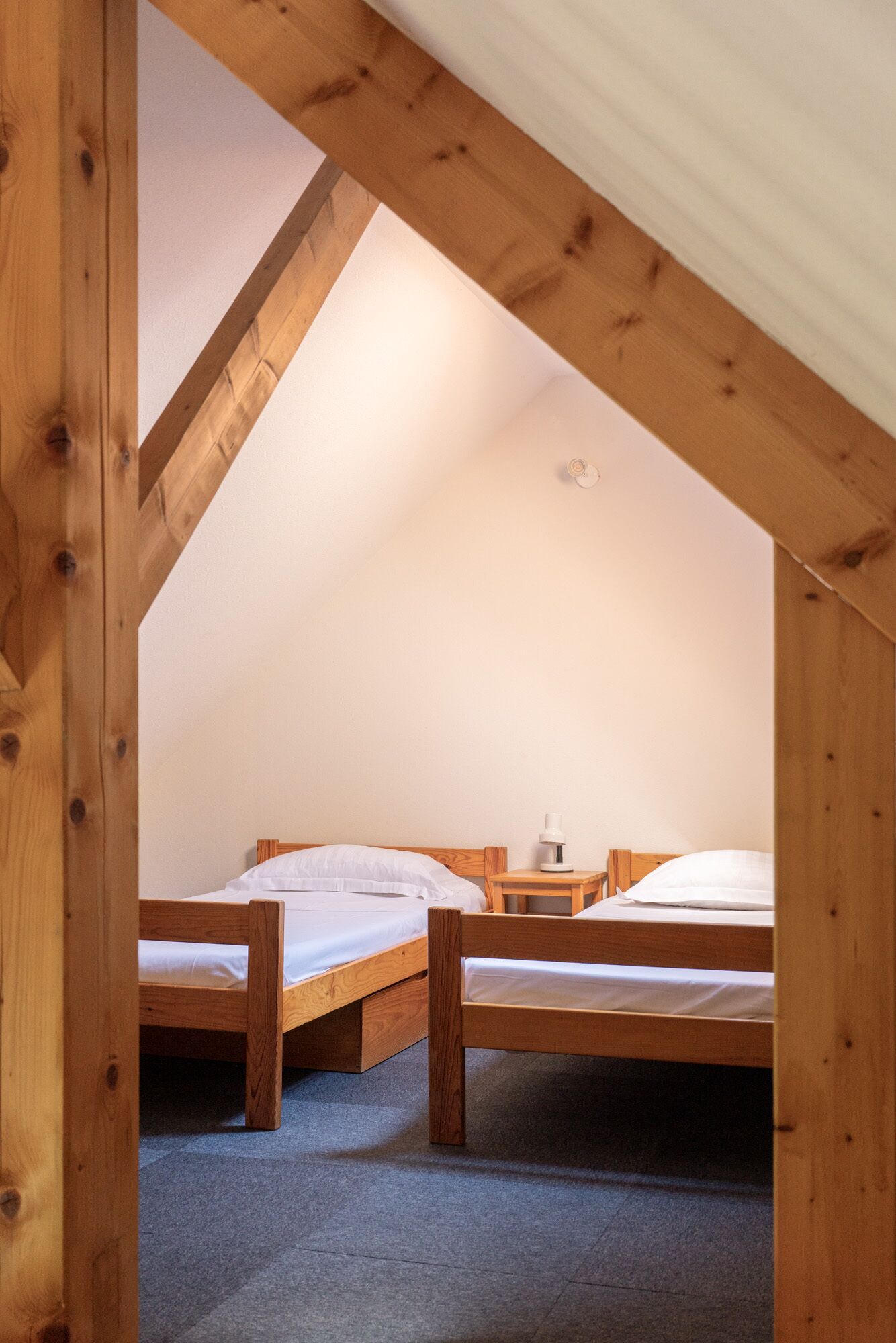 Um quarto pequeno e acolhedor com duas camas de madeira sob um teto inclinado, uma mesa de cabeceira com abajur e piso coberto por carpete.