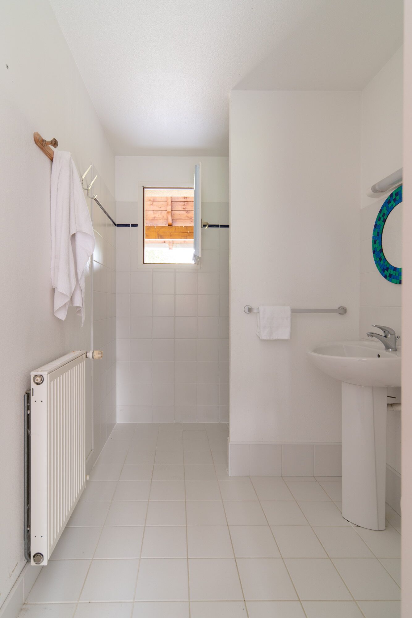 A narrow, white-tiled bathroom with a shower, a white sink, and a towel rack.