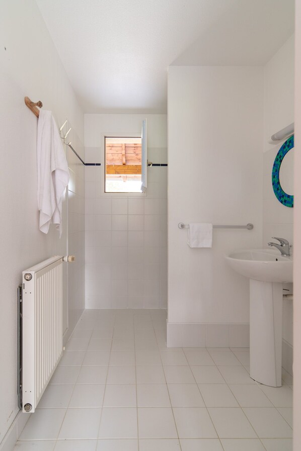 Un baño estrecho con paredes de azulejos blancos, una ducha, un lavamanos blanco y un perchero para toallas.
