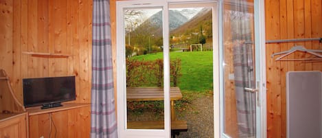 Een kamer met houten wanden, een televisie op een plank, een schuifdeur naar een tuin met een bankje en een hangende kapstok.