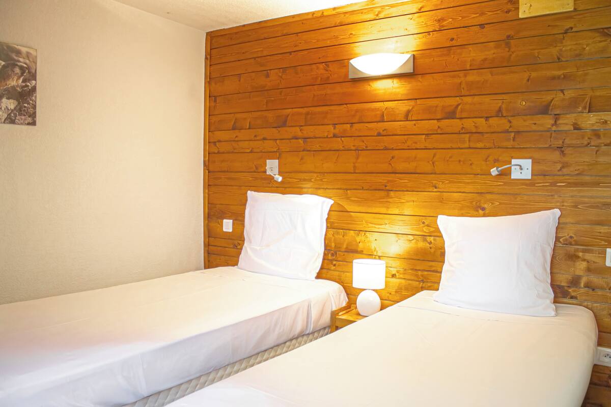 Deux lits simples avec du linge de lit blanc, dans une chambre aux murs lambrissés et dotée d’une petite table de chevet avec une lampe.