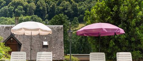 A patio area with four white lounge chairs and two umbrellas, one white and one pink, overlooking a forested hillside.