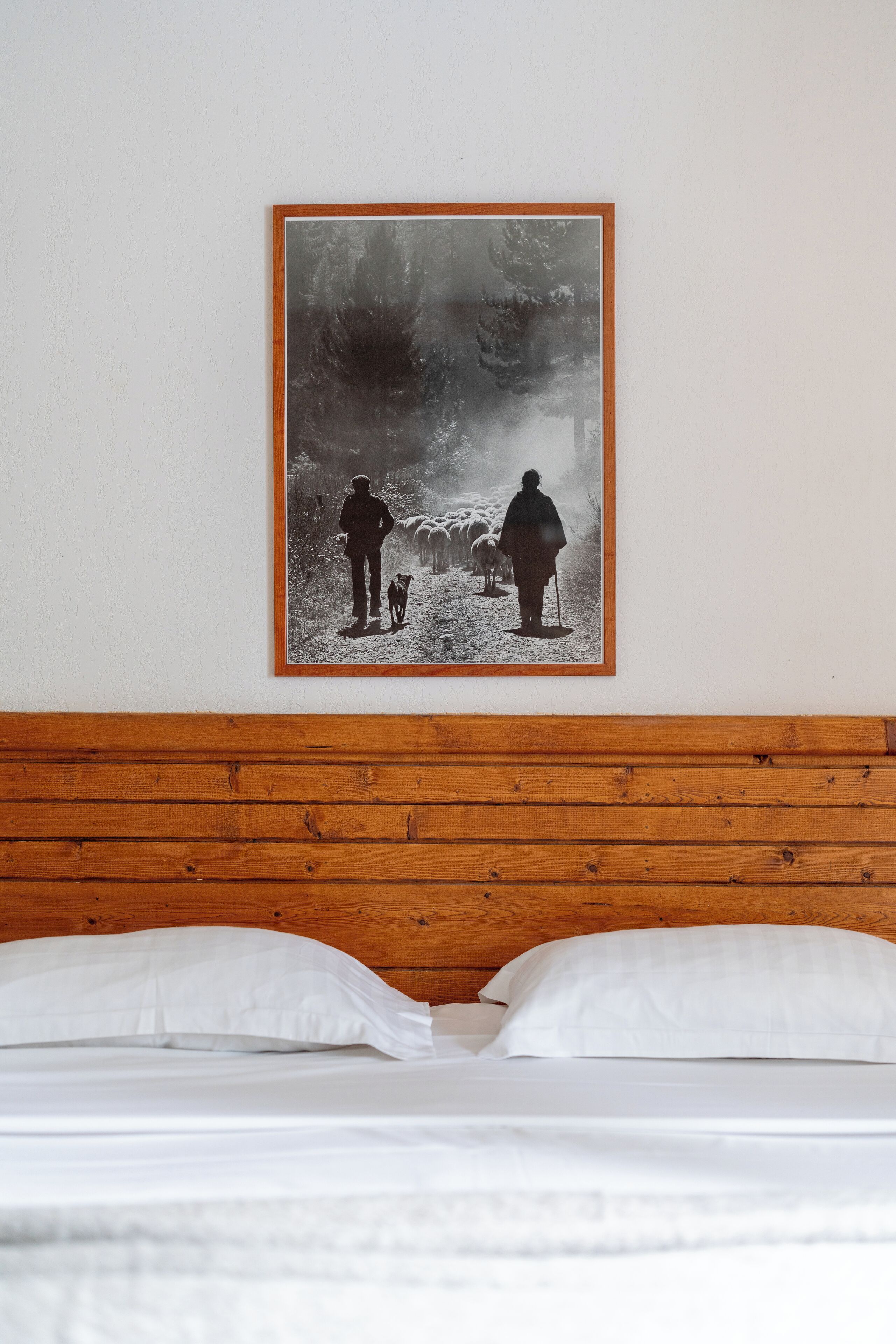 A bedroom with a wooden headboard, white bedding, and a framed picture of a person with sheep on the wall.