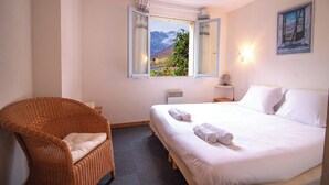 Una habitación de hotel con una cama, una silla de mimbre, una ventana con vista a la montaña y una lámpara fijada a la pared.