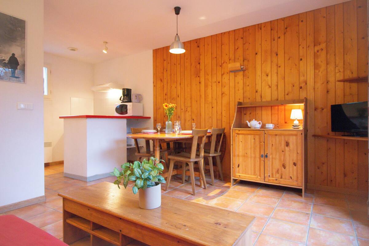 Une cuisine chaleureuse avec des murs en bois, une table à manger avec des chaises, un banc, une plante en pot et un téléviseur posé sur un meuble en bois.