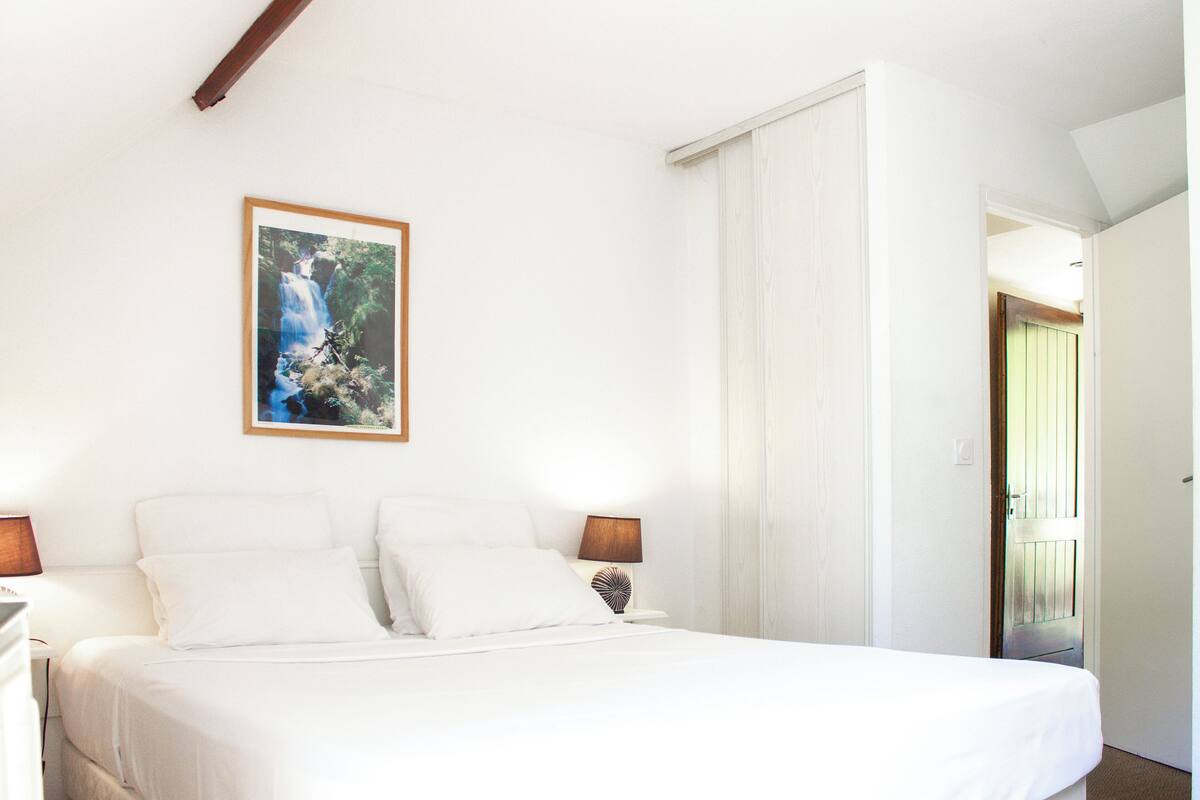 Une chambre à coucher avec un lit, des murs blancs, un tableau encadré représentant une cascade et une porte donnant sur une autre pièce.