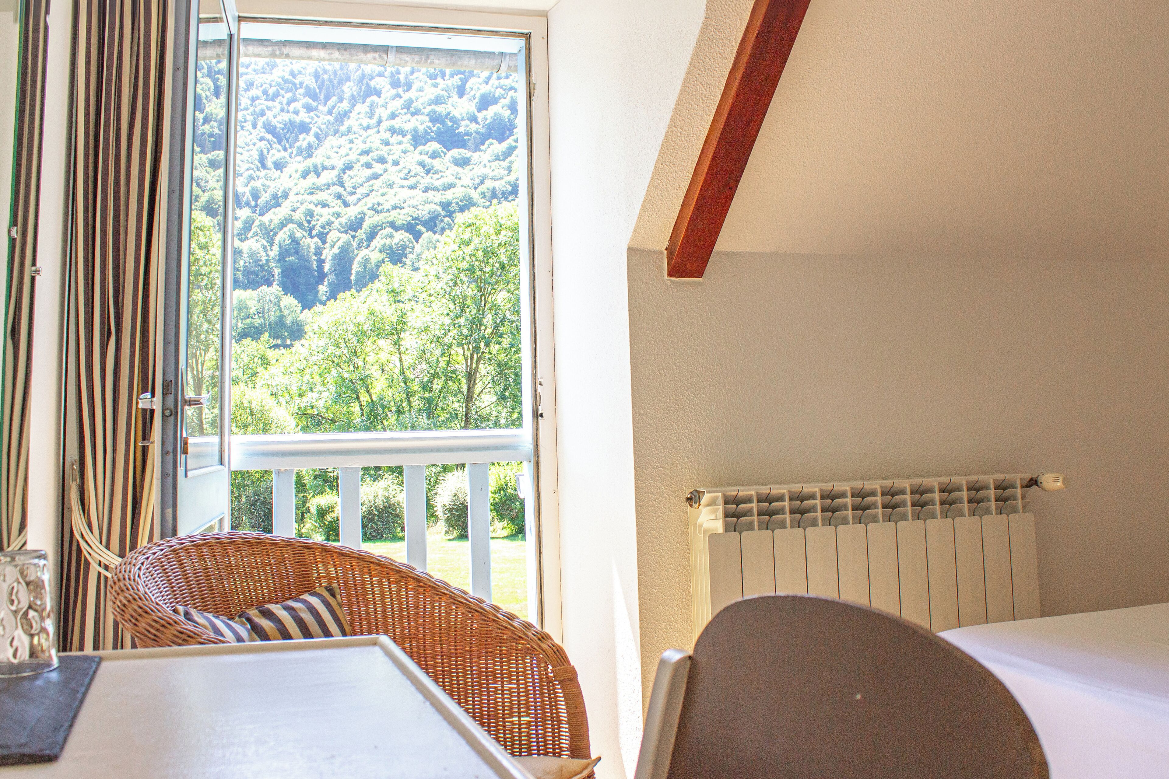 A room with a view of a forest through a window, a wicker chair, a table, and a bed with a white headboard.