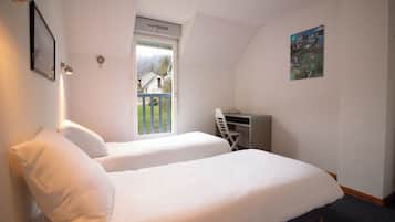 A bedroom with two beds, a desk, and a window with a view of a house and greenery.
