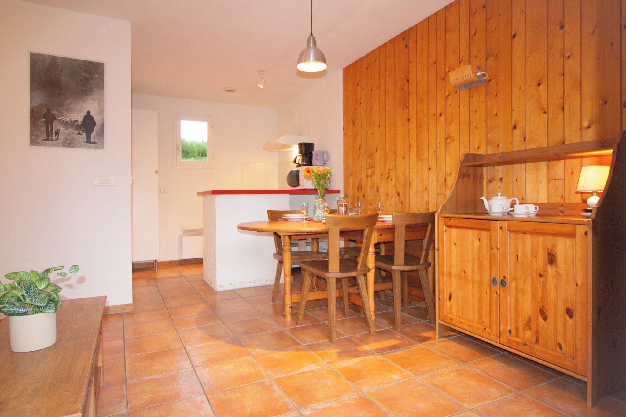 A kitchen with wooden cabinets, a dining table with chairs, a potted plant, and a painting on the wall.