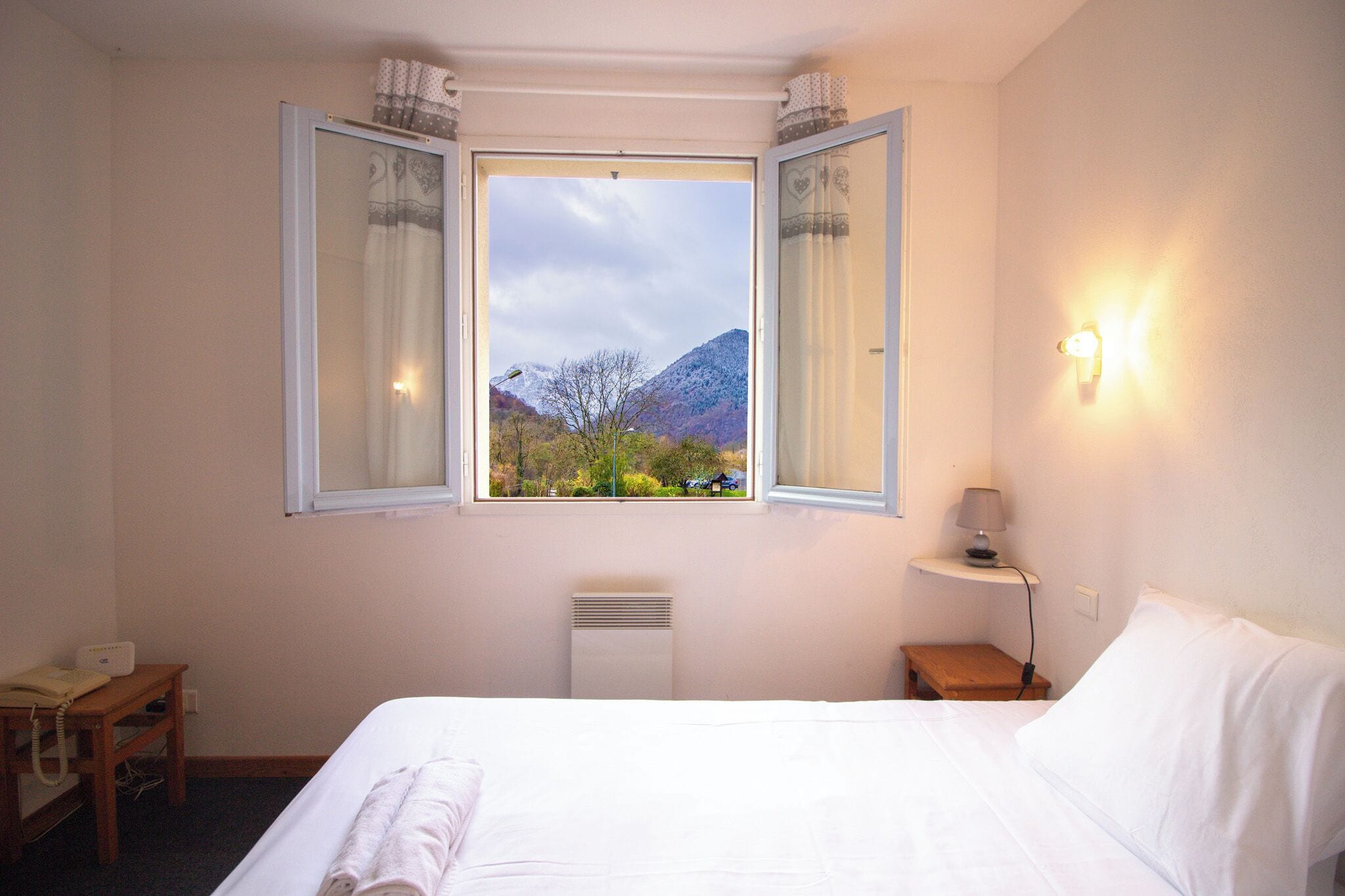 Una habitación de hotel con cama, mesita de noche con lámpara, ventana con cortinas y vistas a las montañas.