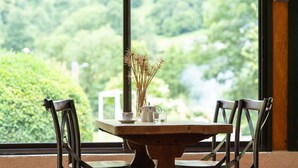 Una mesa de madera con un jarrón de plantas secas, una tetera y una taza, frente a una ventana que ofrece vistas a un paisaje verde.