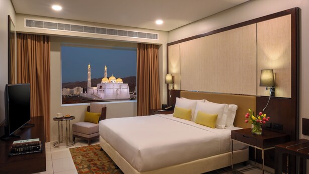 A hotel room with a large bed, a television, a small table, and a view of a cityscape with a prominent building. - Fraser Suites Muscat (Muscat)
