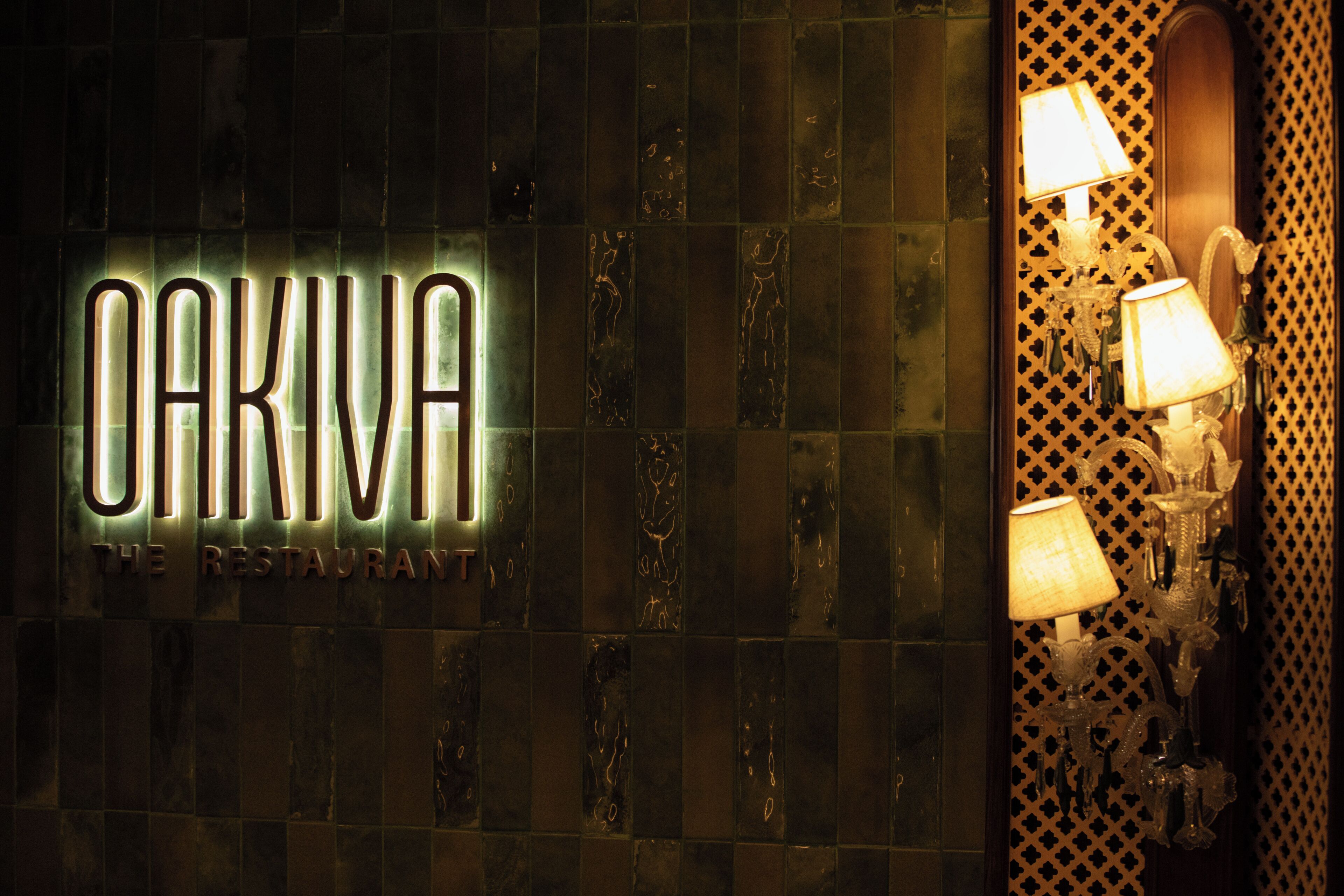 A wall with the illuminated sign 'OAKIVA THE RESTAURANT' and a patterned wall with wall sconces.