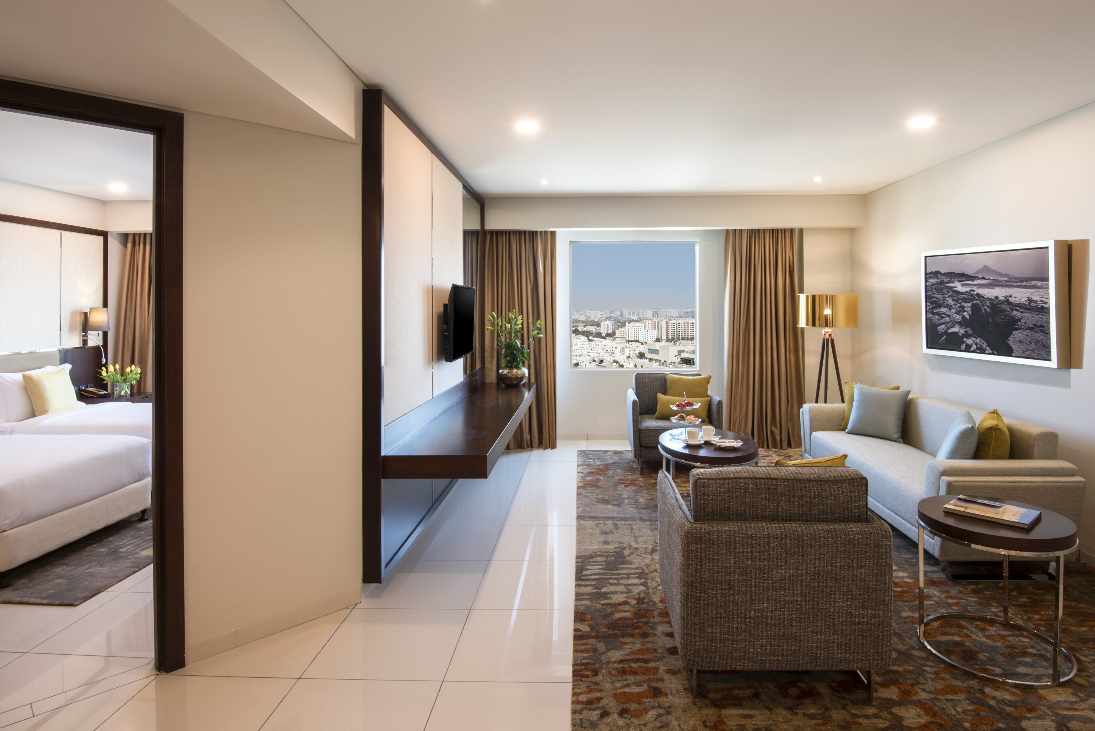 A hotel room with a bed, a seating area with a sofa and armchairs, a small table, a television, and a large window with a city view.