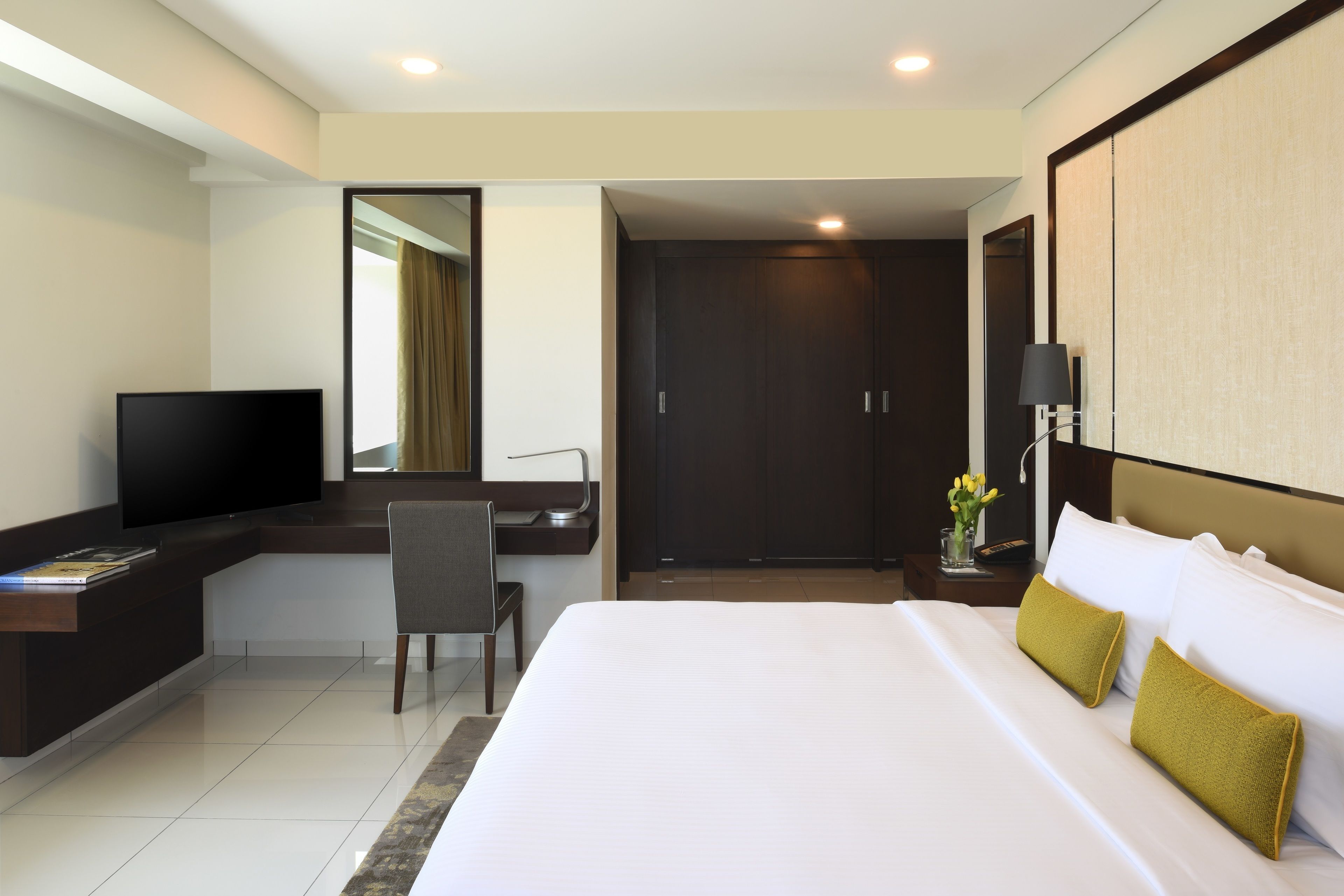 A hotel room with a bed, a desk with a TV, a chair, a nightstand with a lamp, and a door.