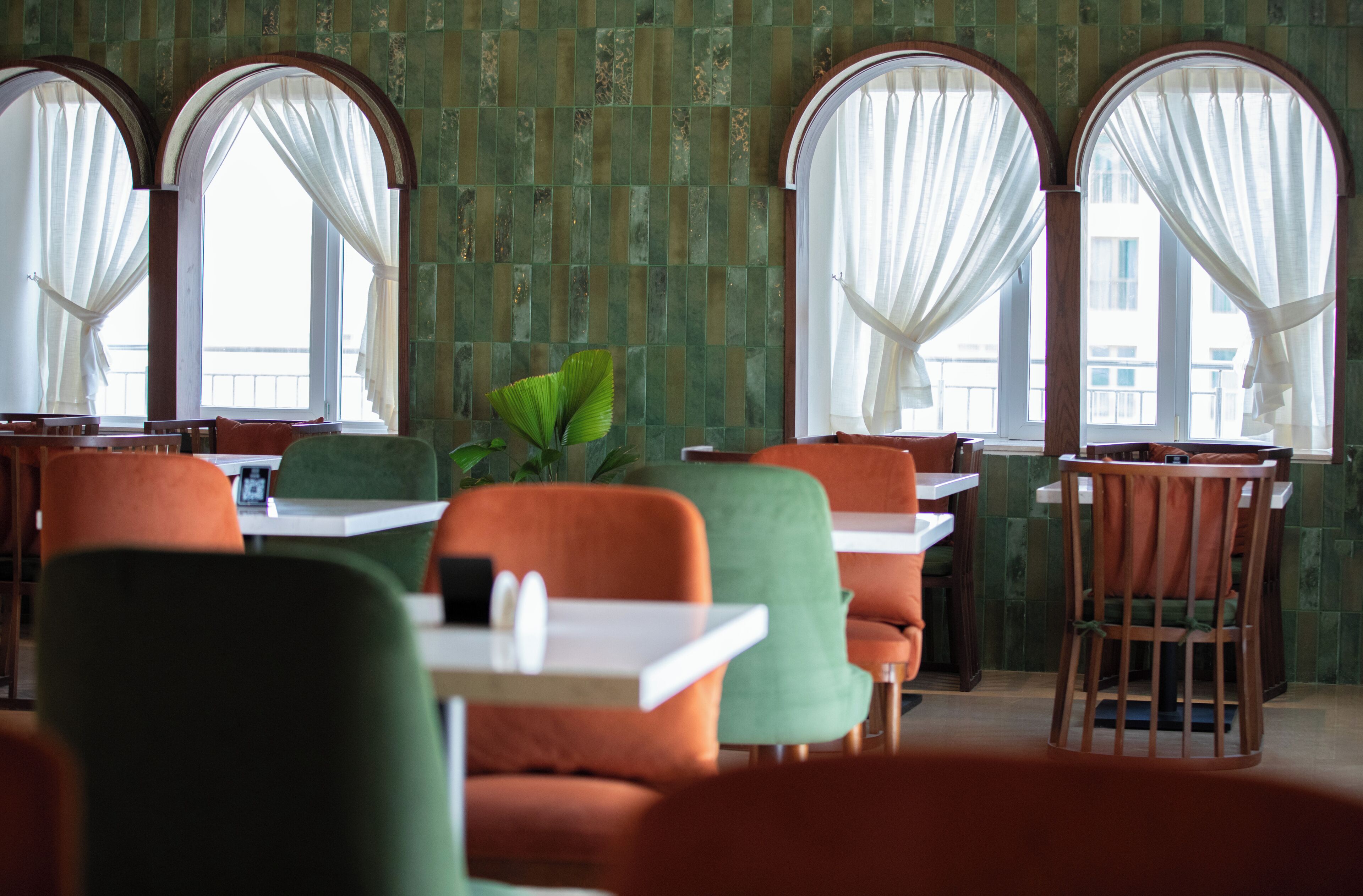 Ein Restaurantinneres mit grünen Fliesenwänden, bogenförmigen Fenstern mit weißen Vorhängen und modernen Möbeln.