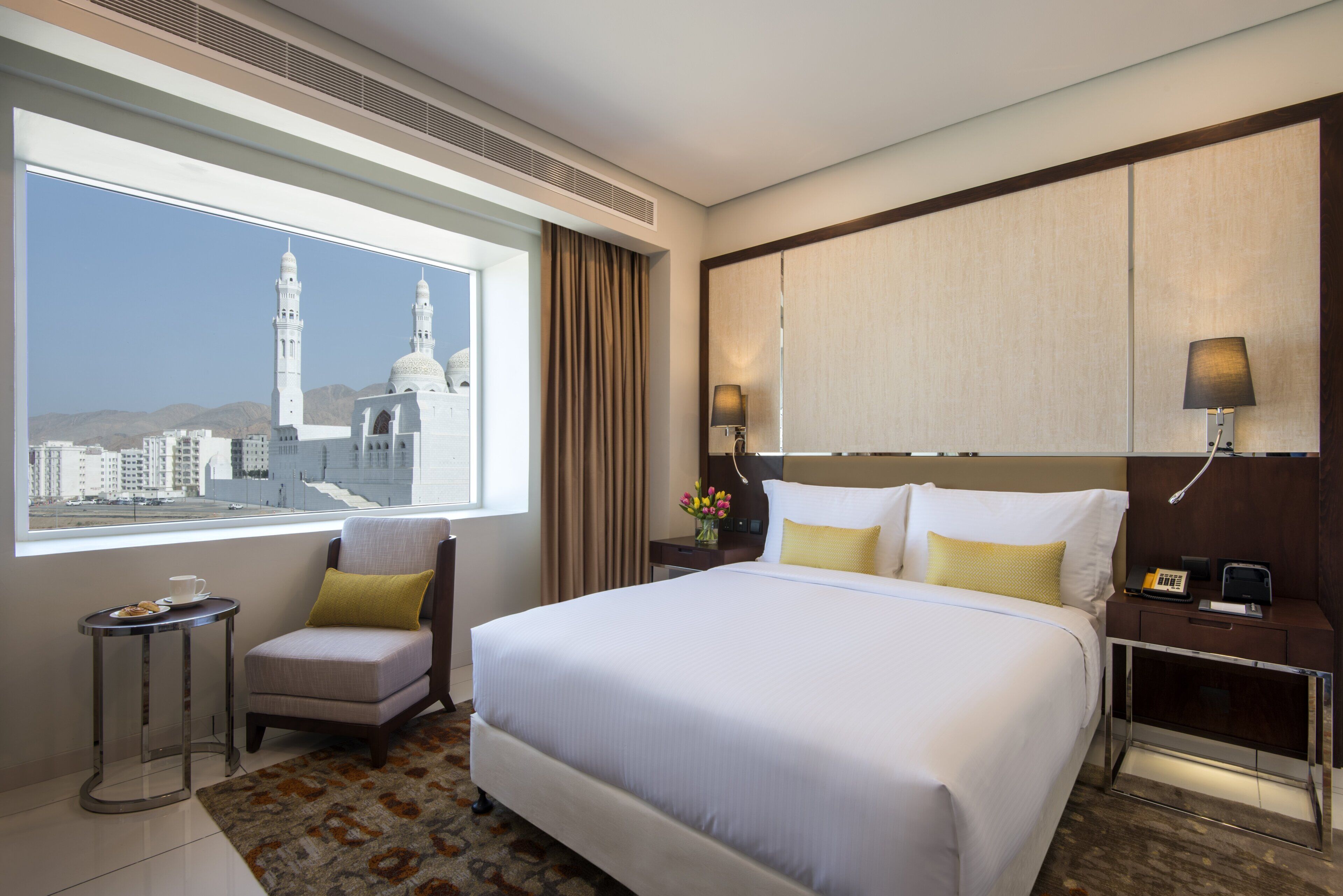 A hotel room with a large bed, a small armchair, a nightstand with a lamp, and a view of a cityscape with a mosque.