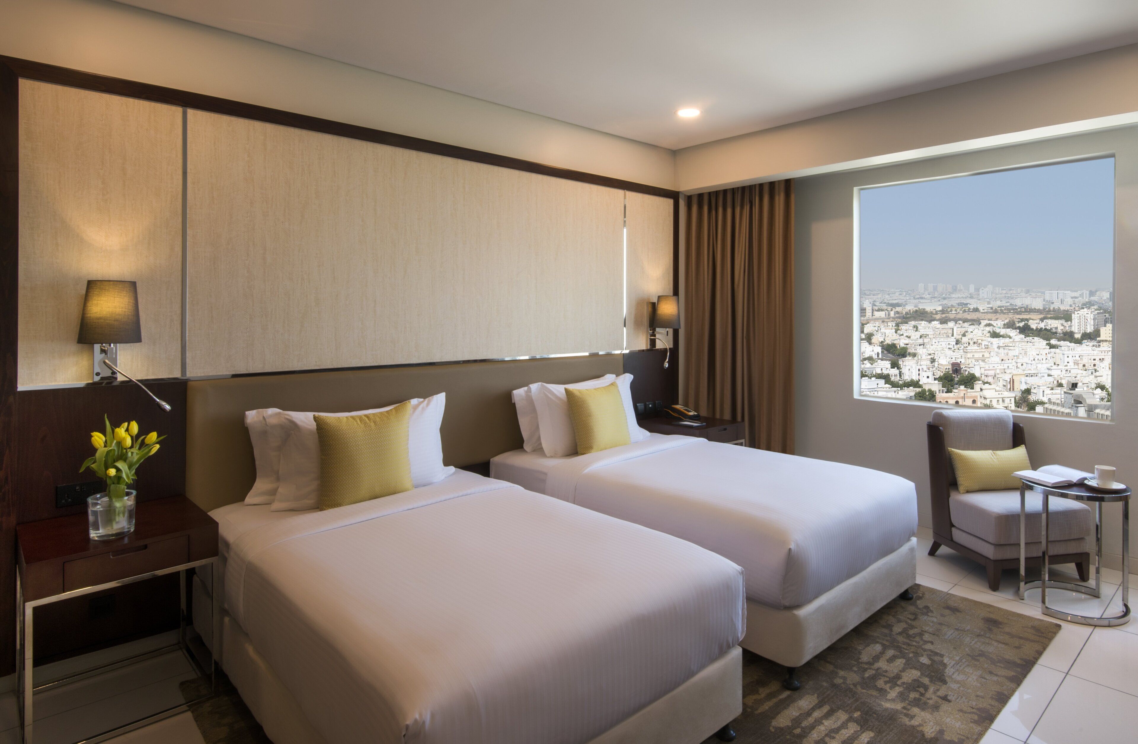 Une chambre d’hôtel avec deux lits, un bureau, une chaise, une petite table et une fenêtre donnant sur la ville.