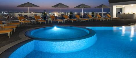 Une piscine entourée de chaises longues et de parasols, offrant une vue sur le paysage urbain au crépuscule.