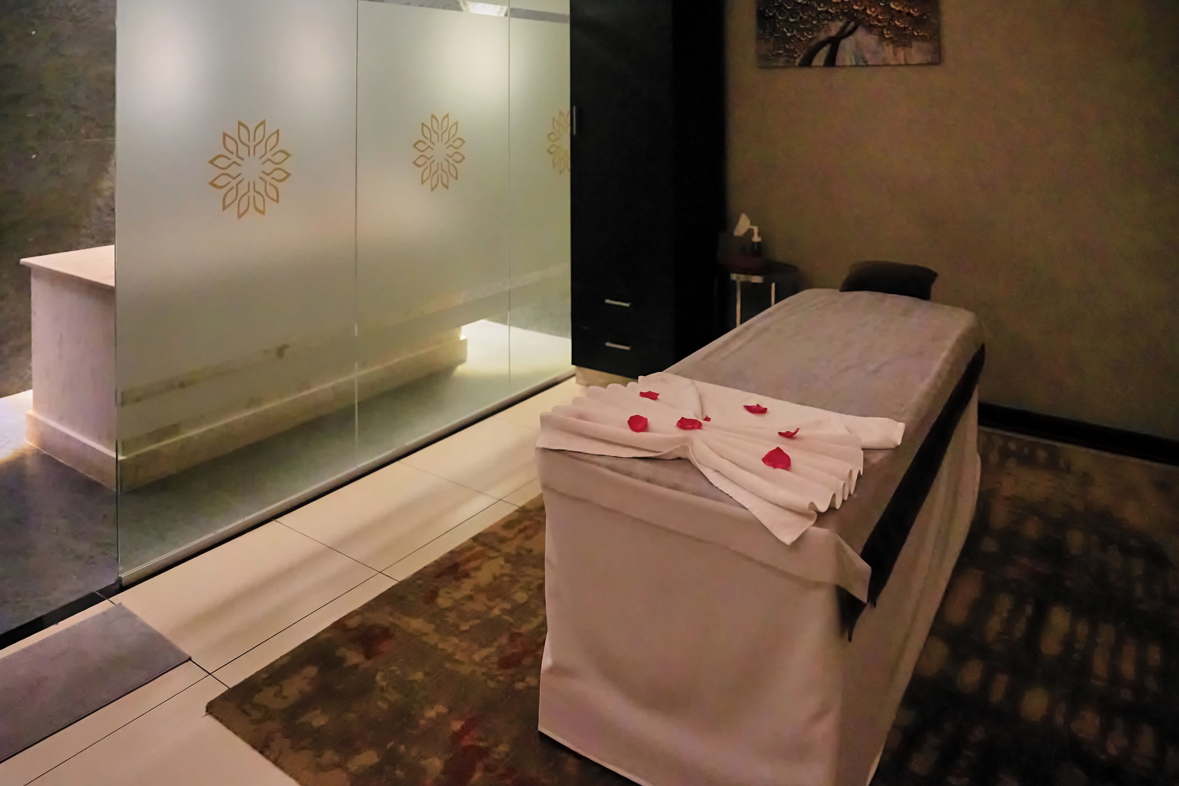 A room with a massage table, a glass partition with a floral design, and a painting on the wall.