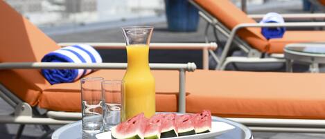 Une table avec un verre de jus d’orange, une tranche de pastèque et deux verres, sur une terrasse sur le toit, agrémentée de chaises longues et de parasols.