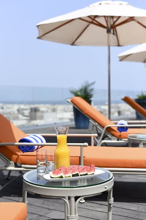 Una mesa con un vaso de jugo de naranja, una tajada de sandía y dos vasos en una terraza en la azotea con sillones y sombrillas.