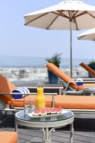 A table with a glass of orange juice, a watermelon slice, and two glasses on a rooftop terrace with lounge chairs and umbrellas.