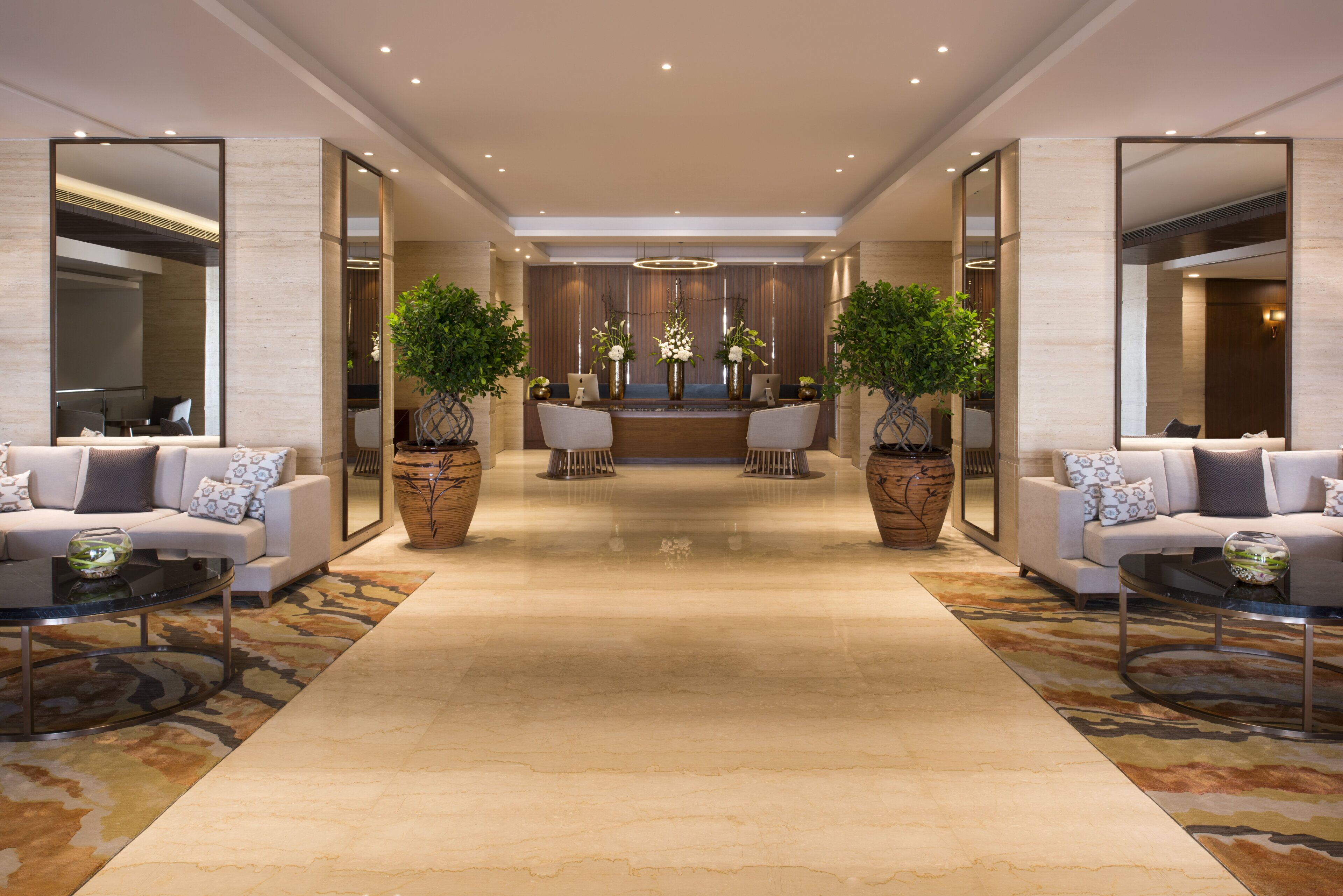 A hotel lobby with a central reception desk, two sofas on either side, and large potted plants.