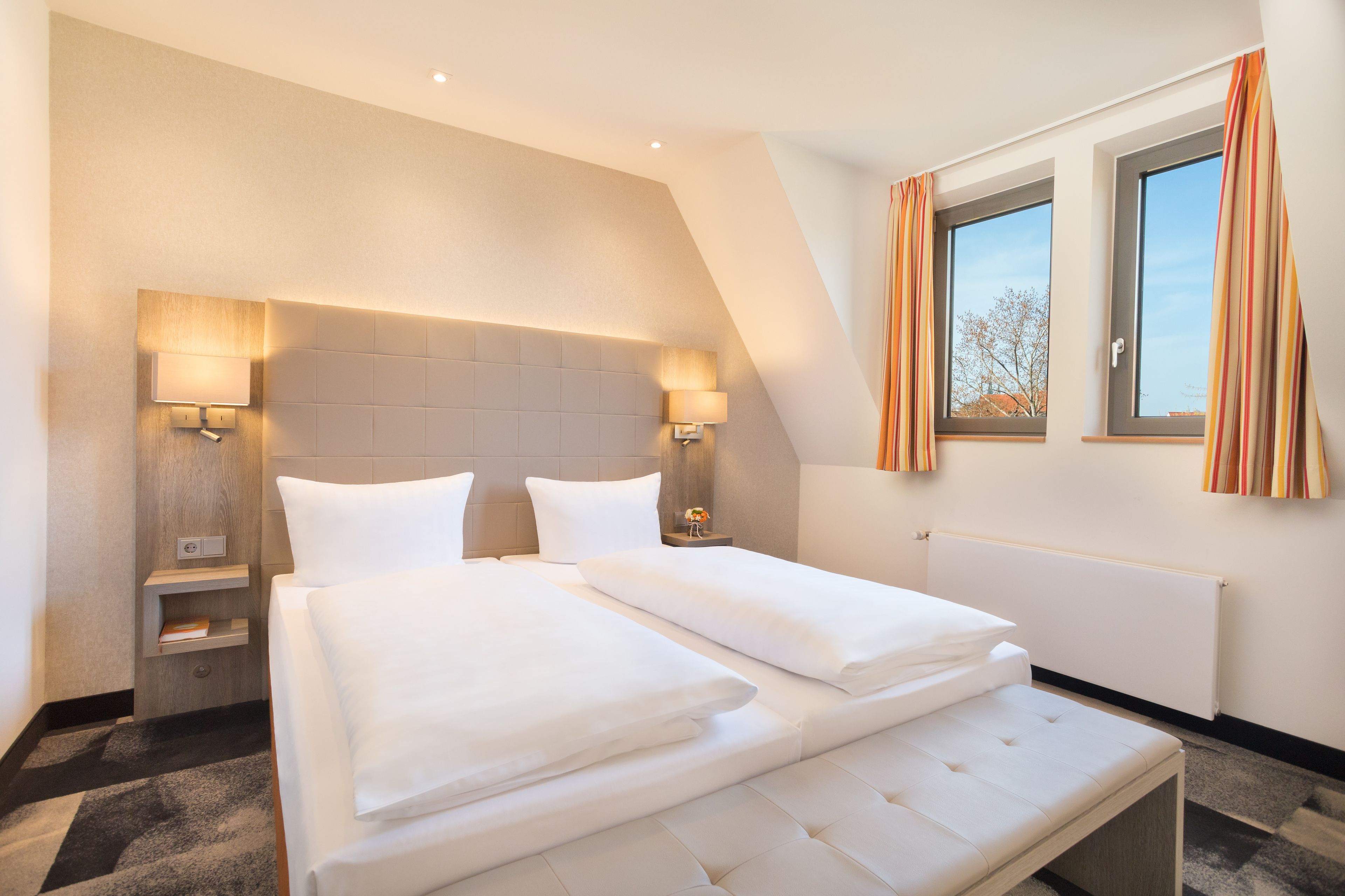 A modern hotel room with a large bed, two bedside tables, a headboard, and a window with curtains.