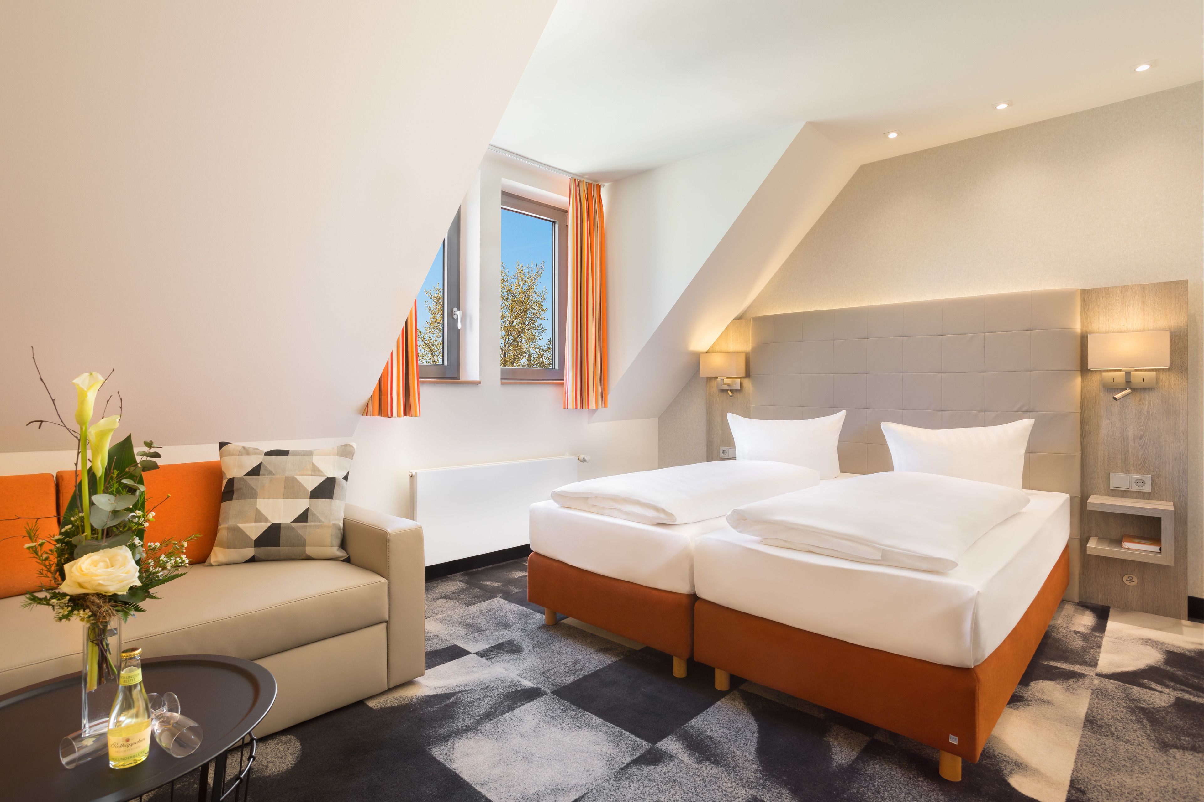 Una habitación de hotel moderna con una cama grande, un sofá, una mesita con un jarrón y un ventanal con cortinas.