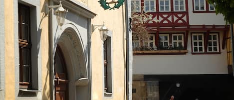 Una calle empedrada con edificios históricos, incluyendo un edificio con un emblema de cisne y una casa tradicional con entramado de madera.